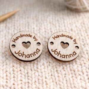 Kuvassa on pyöreä puinen koivuvanerista tehty käsityömerkki, merkin voi kiinnittää omaan käsityöhönsä siinä olevista reiistä, merkissä lukee kauniilla fontilla handmade by Johanna