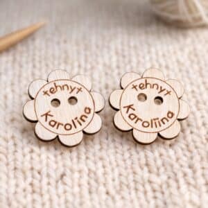 kukan mallinen käsityömerkki, jossa terälehdet kiertävät kehää, keskellä reiät kiinnitystä varten, merkki on personoitu tekstillä "tehnyt Karoliina"