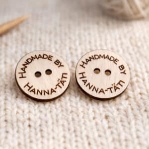 pyöreä käsityömerkki Handmade by Hanna-täti. Merkki on personoitava ja teksti on kirjoitettu suuraakkosin. Merkin keskellä on kaksi reikää kiinnitystä varten.