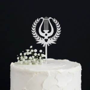 Valkoinen puinen ylioppilaskakkukoriste lyyra-symbolilla ja lehväseppeleellä valkoisen täytekakun päällä.