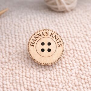 puinen pylreä käsityömerkki vaalan neulotun materiaalin päällä, merkkiä kehystää katkoviiva, merkissä neljä reikää kiinnitystä varten, merkki personoitavissa omalla tekstillä