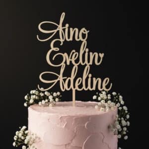 Lähikuva kakkukoristeesta, jossa lukee nimi "Olivia". Puinen kakkukoriste on asetettu vaaleanpunaisilla kukilla ja mustikoilla koristellun valkoisen täytekakun päälle.